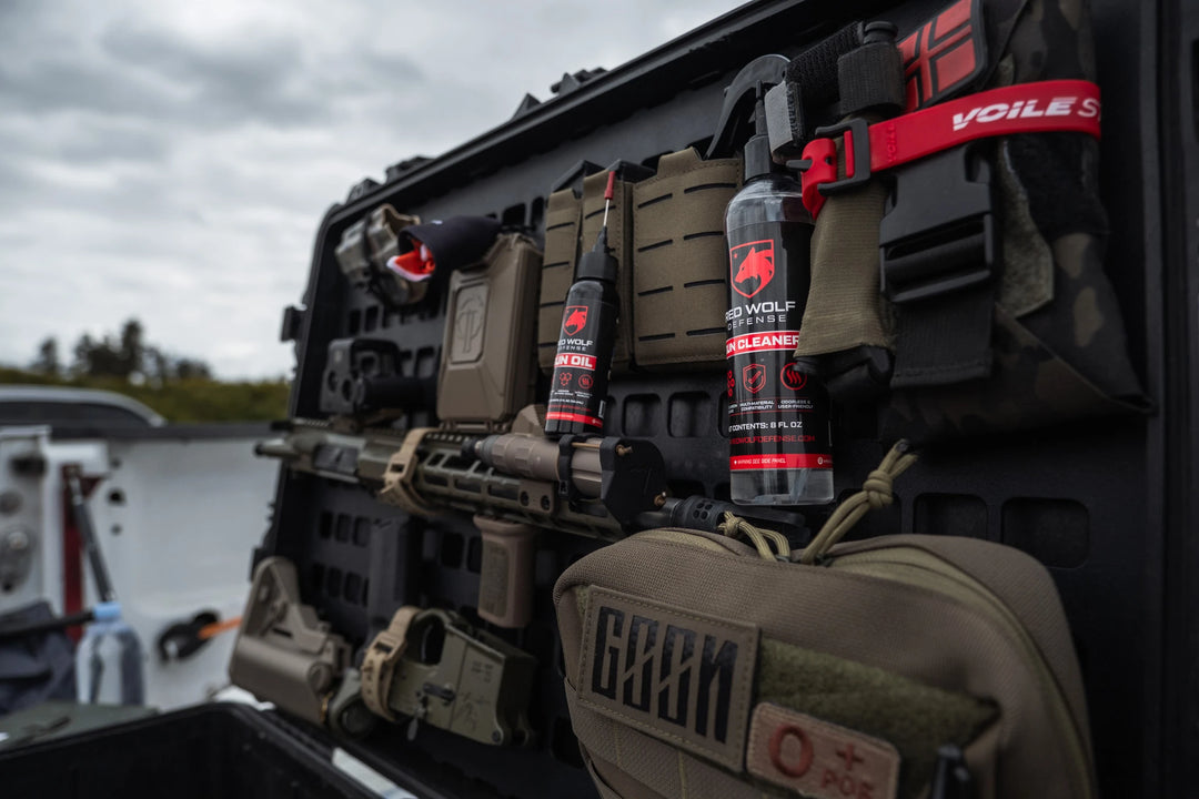 Red Wolf Defense gun cleaning products including gun oil and gun cleaner displayed on a tactical gear setup, with various military-style accessories and equipment in the background.