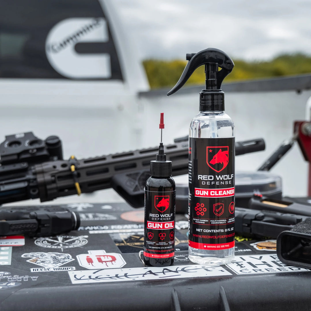 Red Wolf Defense gun cleaner and gun oil bottles placed on a tactical surface with firearms and gear in the background.